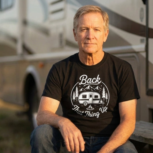A man wearing a black graphic t-shirt with a white 'Back to the Thing Up' RV design, sitting in front of a large recreational vehicle outdoors.
