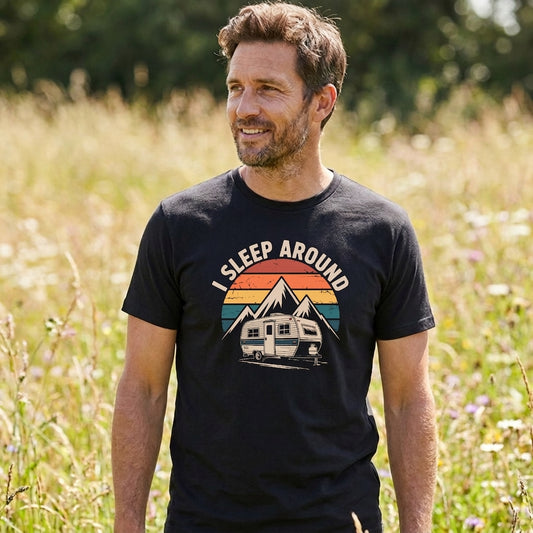 A man wearing a black graphic t-shirt with a retro camping trailer design and the text 'I SLEEP AROUND' standing in a sunlit grassy field.