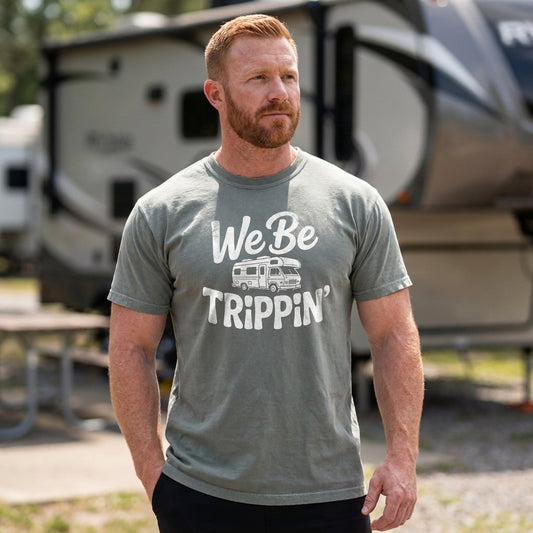 A man wearing a heather gray t-shirt with a white graphic that reads 'We Be TRIPPIN'' and an RV illustration, standing outdoors in front of a blurred travel trailer.