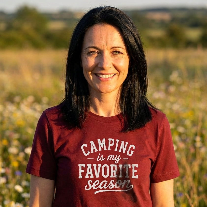 A woman with dark hair stands in a sunlit field wearing a burgundy t-shirt with the text 'CAMPING is my FAVORITE season' in white and light red lettering.