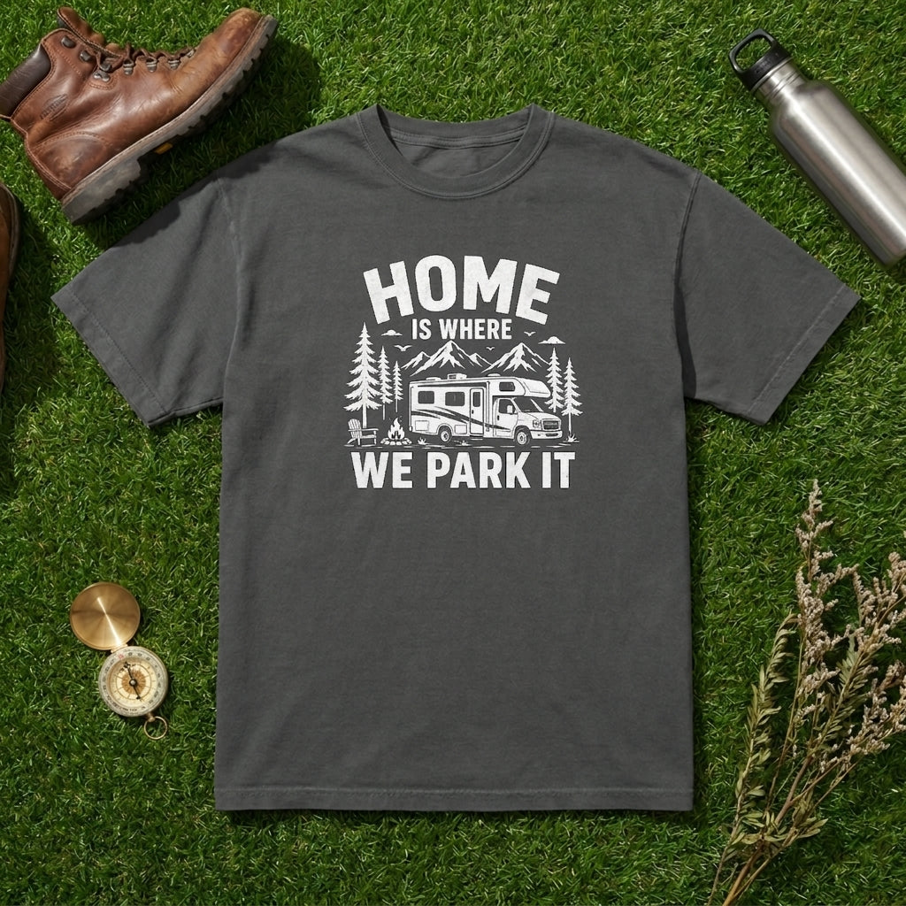 A dark gray crewneck t-shirt with a white graphic print reading 'HOME IS WHERE WE PARK IT' with a camper and trees, laid flat on green grass with outdoor props including a brown boot, metal water bottle, compass, and dried flowers.