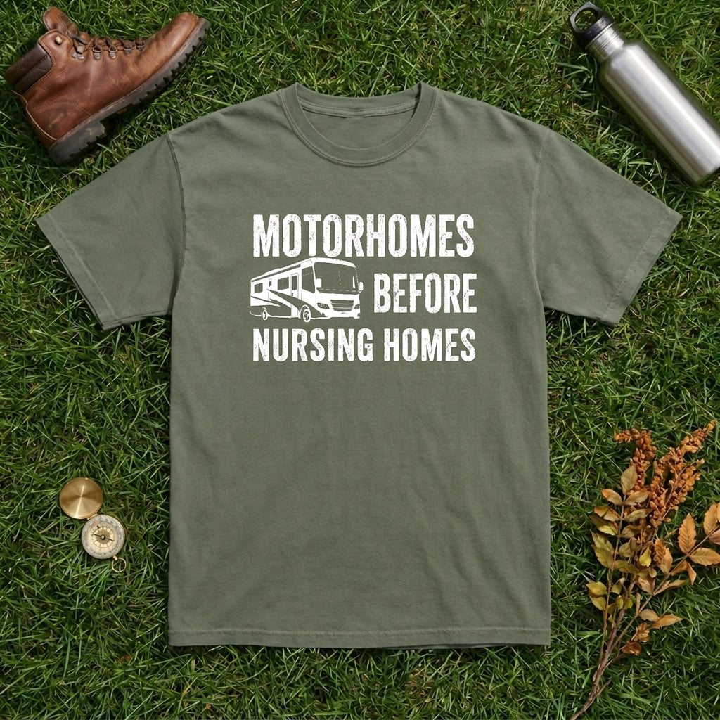 An olive green crewneck t-shirt with a white graphic print reading 'MOTORHOMES BEFORE NURSING HOMES' with an RV illustration, laid flat on green grass with a brown boot, metal water bottle, and dried leaves as props.