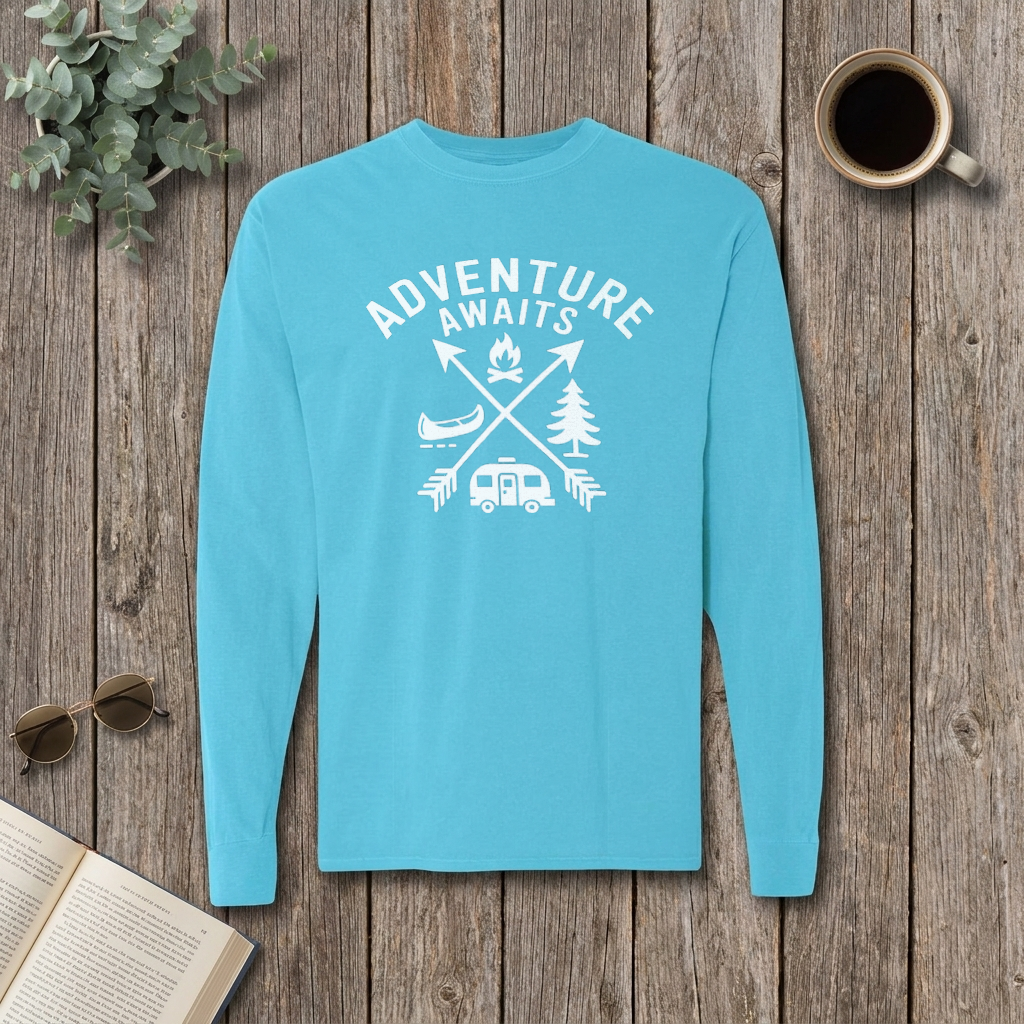 Flat lay of a light blue Adventure Awaits long sleeve camping shirt on a wood table with coffee and sunglasses