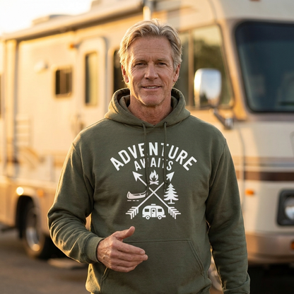 Smiling man in an olive green Adventure Awaits camping hoodie standing in front of a motorhome