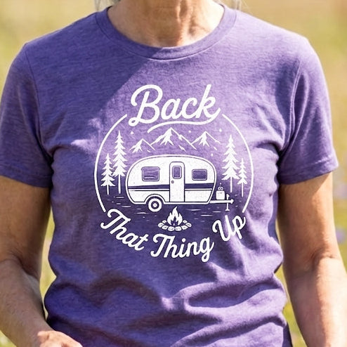 A smiling older woman standing in a sunlit field of wildflowers, wearing a heather purple graphic T-shirt with a white camping camper and trees print that reads 'Back That Thing Up.'.
