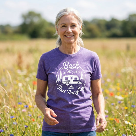 A smiling older woman standing in a sunlit field of wildflowers, wearing a heather purple graphic T-shirt with a white camping camper and trees print that reads 'Back That Thing Up.'.