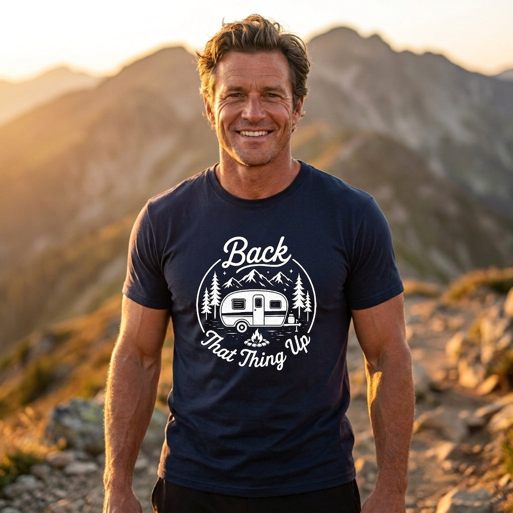 A smiling man standing outdoors in front of mountains at sunset, wearing a navy blue graphic T-shirt with a white camping trailer and trees design and the words 'Back That Thing Up.'.