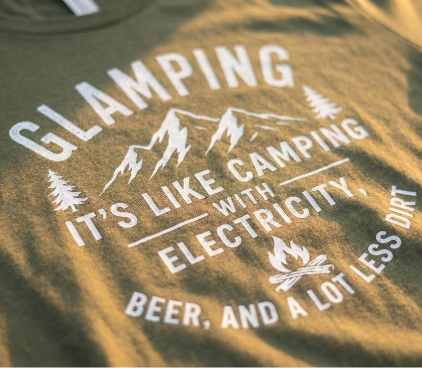 Close-up of a military green t-shirt featuring a white graphic print that reads 'GLAMPING IT'S LIKE CAMPING WITH ELECTRICITY, BEER, AND A LOT LESS DIRT' with mountain and campfire illustrations.