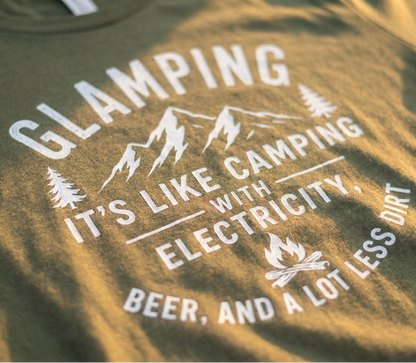 Close-up of a military green t-shirt featuring a white graphic print that reads 'GLAMPING IT'S LIKE CAMPING WITH ELECTRICITY, BEER, AND A LOT LESS DIRT' with mountain and campfire illustrations.