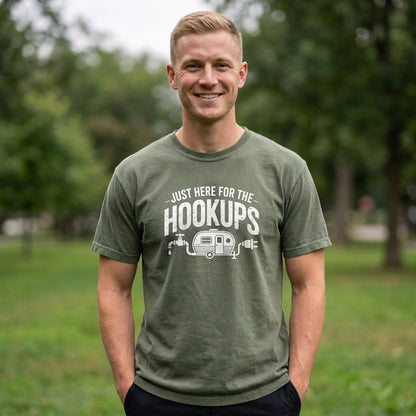 A smiling man wearing a military green t-shirt with a white graphic that reads 'JUST HERE FOR THE HOOKUPS' and features a small trailer illustration, standing in a park with trees in the background.