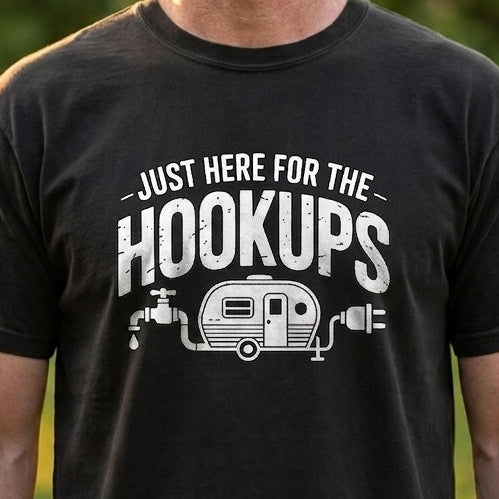 A man wearing a black graphic t-shirt with a white print that reads 'JUST HERE FOR THE HOOKUPS' and features a small trailer illustration. The background is a blurred outdoor park scene with green grass and trees.