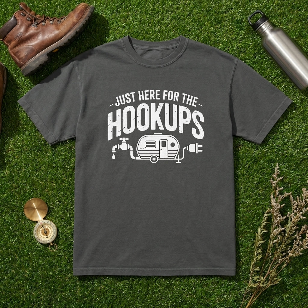 A dark gray crewneck t-shirt with a white graphic reading 'JUST HERE FOR THE HOOKUPS' and a camper trailer illustration, laid flat on artificial green grass with a brown leather boot, metal water bottle, compass, and dried flowers as props.