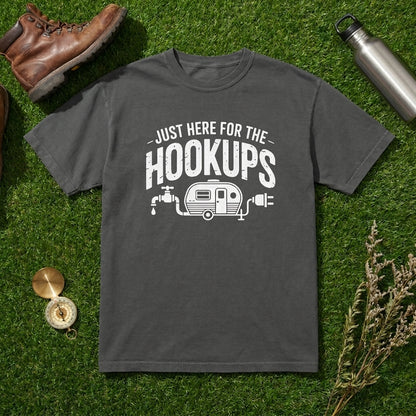 A dark gray crewneck t-shirt with a white graphic reading 'JUST HERE FOR THE HOOKUPS' and a camper trailer illustration, laid flat on artificial green grass with a brown leather boot, metal water bottle, compass, and dried flowers as props.