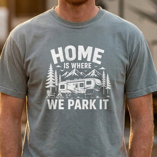 A man wearing a gray graphic t-shirt with the text 'HOME IS WHERE WE PARK IT' and a camping RV design, standing in front of a blurred RV at sunset.