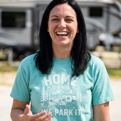 A woman smiling outdoors wearing a light aqua t-shirt with a white graphic that reads 'We Be TRIPPIN'' and shows a campervan illustration. A blurred RV is visible in the background.