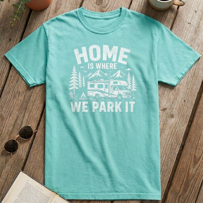 A mint green short-sleeve t-shirt with a white graphic print reading 'HOME IS WHERE WE PARK IT' and showing mountains, trees, and an RV, laid flat on a rustic wooden surface with sunglasses, an open book, and a mug nearby.