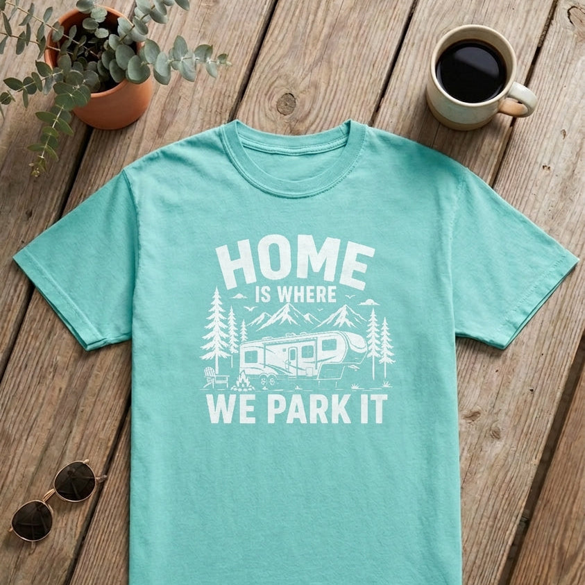 A mint green crewneck t-shirt with a white graphic print reading 'HOME IS WHERE WE PARK IT' with a camper and trees, laid flat on a rustic wooden table with a coffee mug, sunglasses, an open book, and a small plant.
