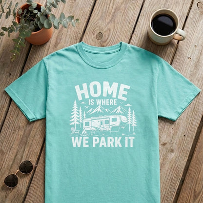 A mint green crewneck t-shirt with a white graphic print reading 'HOME IS WHERE WE PARK IT' with a camper and trees, laid flat on a rustic wooden table with a coffee mug, sunglasses, an open book, and a small plant.