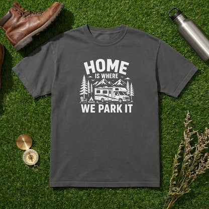 A dark gray crewneck t-shirt with a white graphic print reading 'HOME IS WHERE WE PARK IT' with a camper and trees, laid flat on green grass with outdoor props including a brown boot, metal water bottle, compass, and dried flowers.