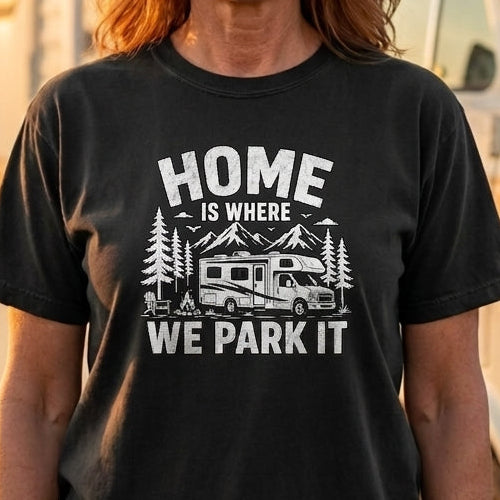 A woman stands outdoors in front of a vintage camper van, wearing a black graphic T-shirt with the white text 'HOME IS WHERE WE PARK IT' and a camping illustration. The shirt is the product being shown, but because a person and real environment are present, the subject_type is Lifestyle.