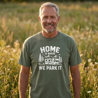 A smiling middle-aged man standing in a sunlit grassy field, wearing an olive green graphic T-shirt with the white printed text 'HOME IS WHERE WE PARK IT' and a camping RV and trees design.