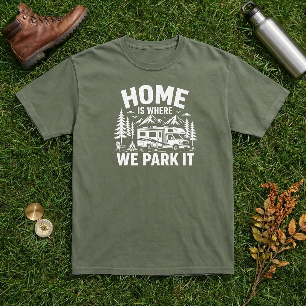 An olive green crewneck t-shirt with a white graphic print reading 'HOME IS WHERE WE PARK IT' with a camper and trees, laid flat on green grass with a brown leather boot, a silver water bottle, and small autumn leaves as props.