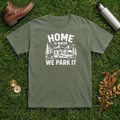 An olive green crewneck t-shirt with a white graphic print reading 'HOME IS WHERE WE PARK IT' with a camper and trees, laid flat on green grass with a brown leather boot, a silver water bottle, and small autumn leaves as props.