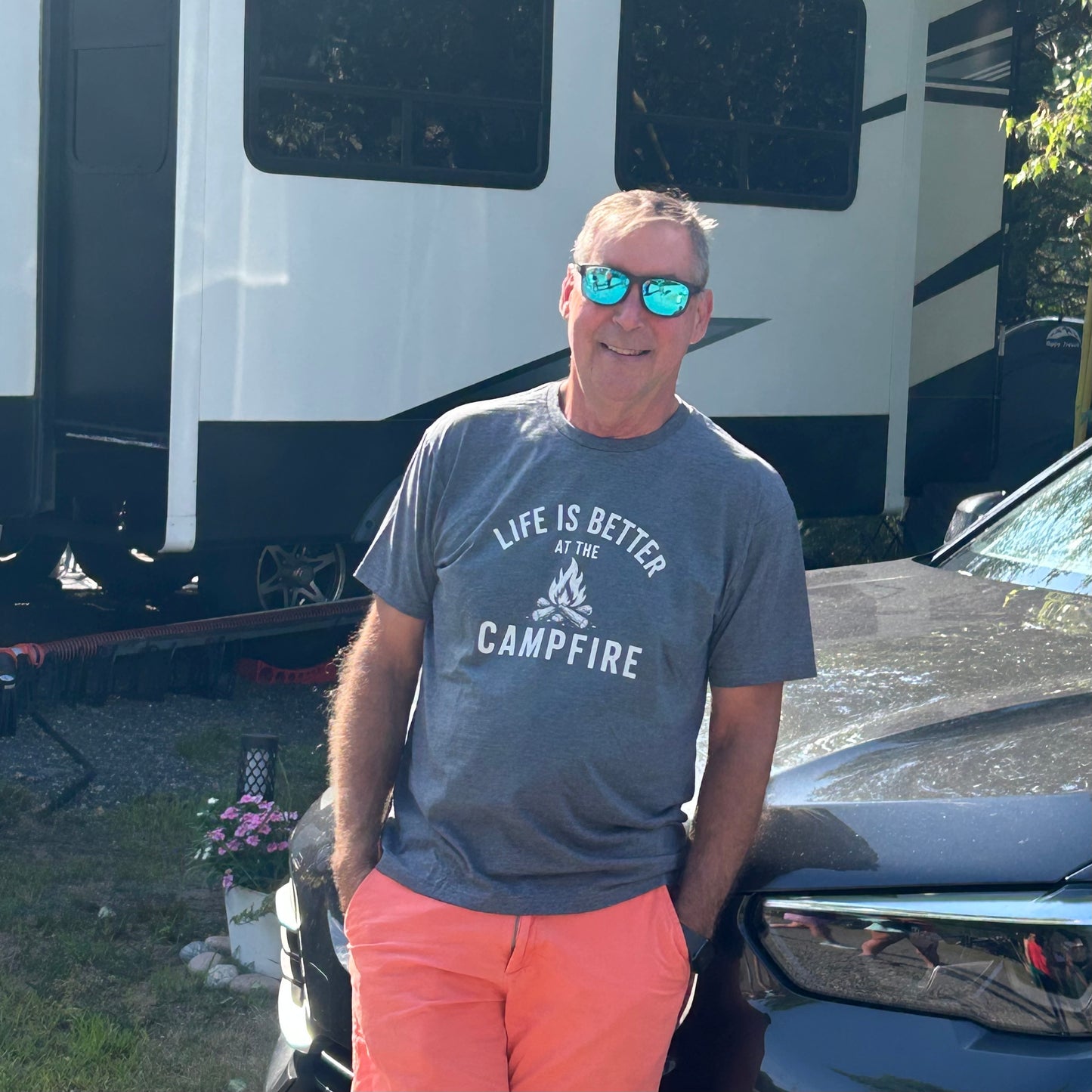A man wearing a gray graphic t-shirt that reads 'LIFE IS BETTER AT THE CAMPFIRE' and bright orange pants, standing outdoors in front of an RV and a dark car.