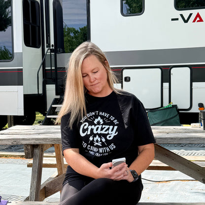 A woman sits on a wooden picnic table in front of an RV, wearing a black graphic T-shirt that reads 'Crazy To Camp With Us' and black leggings.