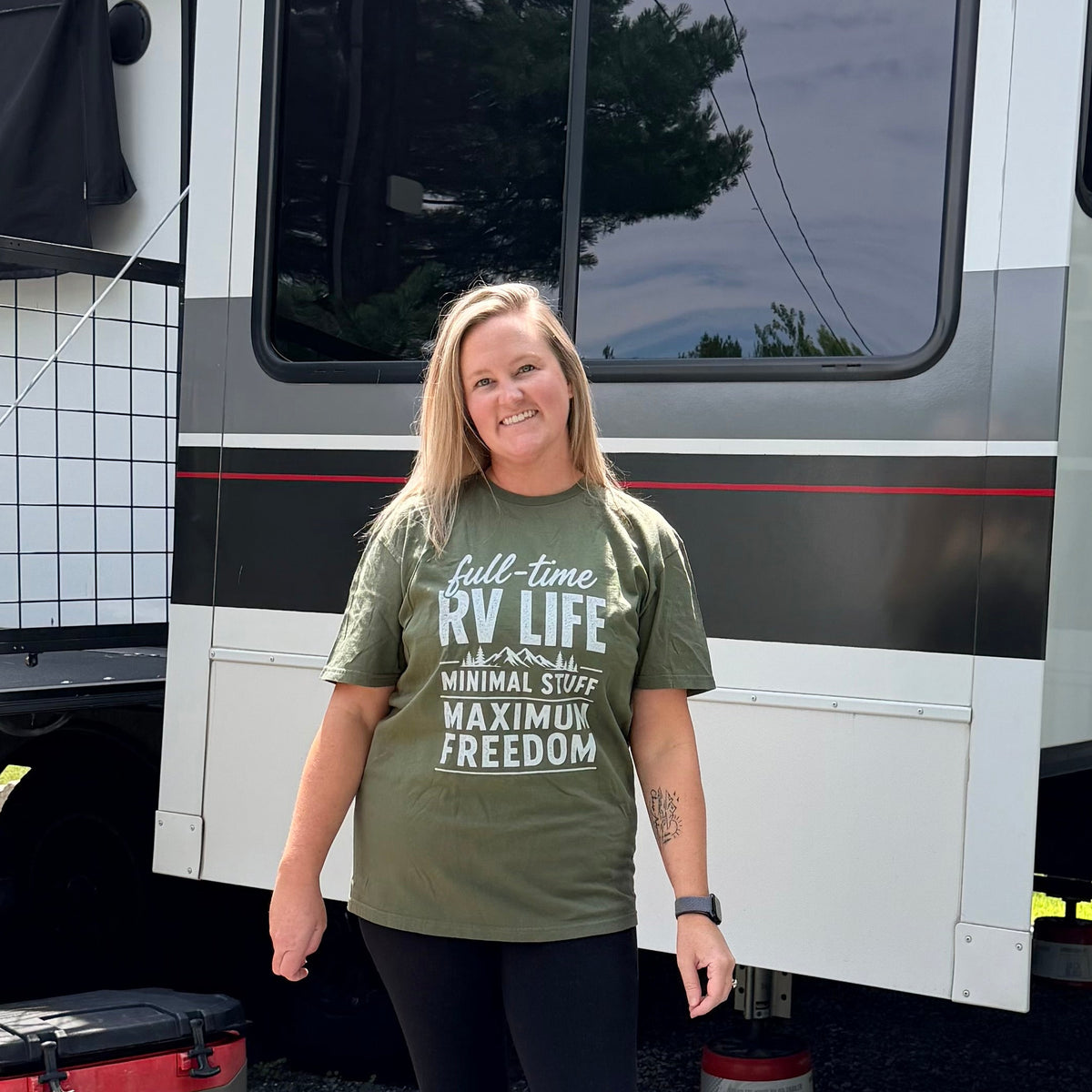 Woman in an olive green full-time RV life t-shirt and black pants standing in front of a large white and gray RV.