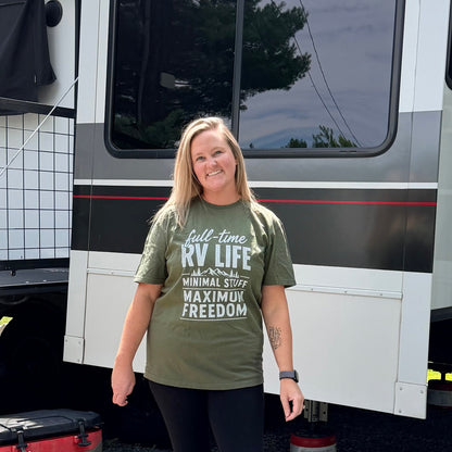 Woman in an olive green full-time RV life t-shirt and black pants standing in front of a large white and gray RV.