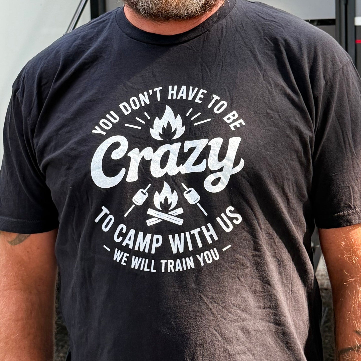 A man wearing a dark charcoal gray graphic t-shirt with a white 'Crazy Camp With Us' print, standing in front of a white RV. The t-shirt features a graphic of a campfire with the text 'YOU DON'T HAVE TO BE Crazy TO CAMP WITH US' and 'WE WILL TRAIN YOU'.