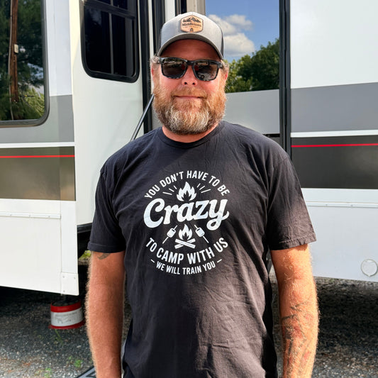 A man wearing a dark charcoal gray graphic t-shirt with a white 'Crazy Camp With Us' print, standing in front of a white RV. The t-shirt features a graphic of a campfire with the text 'YOU DON'T HAVE TO BE Crazy TO CAMP WITH US' and 'WE WILL TRAIN YOU'.