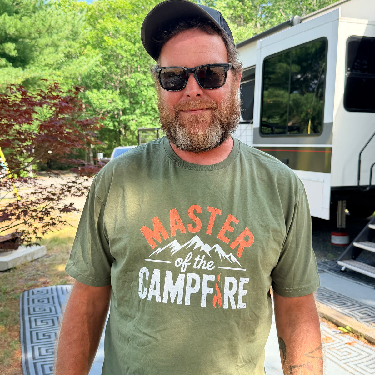 Man in a green Master of the Campfire t-shirt standing outdoors at a campsite.