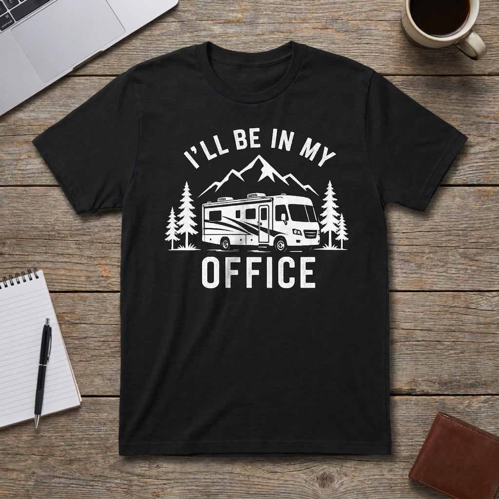 A black short-sleeve graphic t-shirt laid flat on a rustic wooden table, featuring a white printed design of an RV, mountains, and pine trees with the text 'I'LL BE IN MY OFFICE'. The shirt is styled with a laptop corner, a coffee mug, a notebook, a pen, and a brown leather wallet.