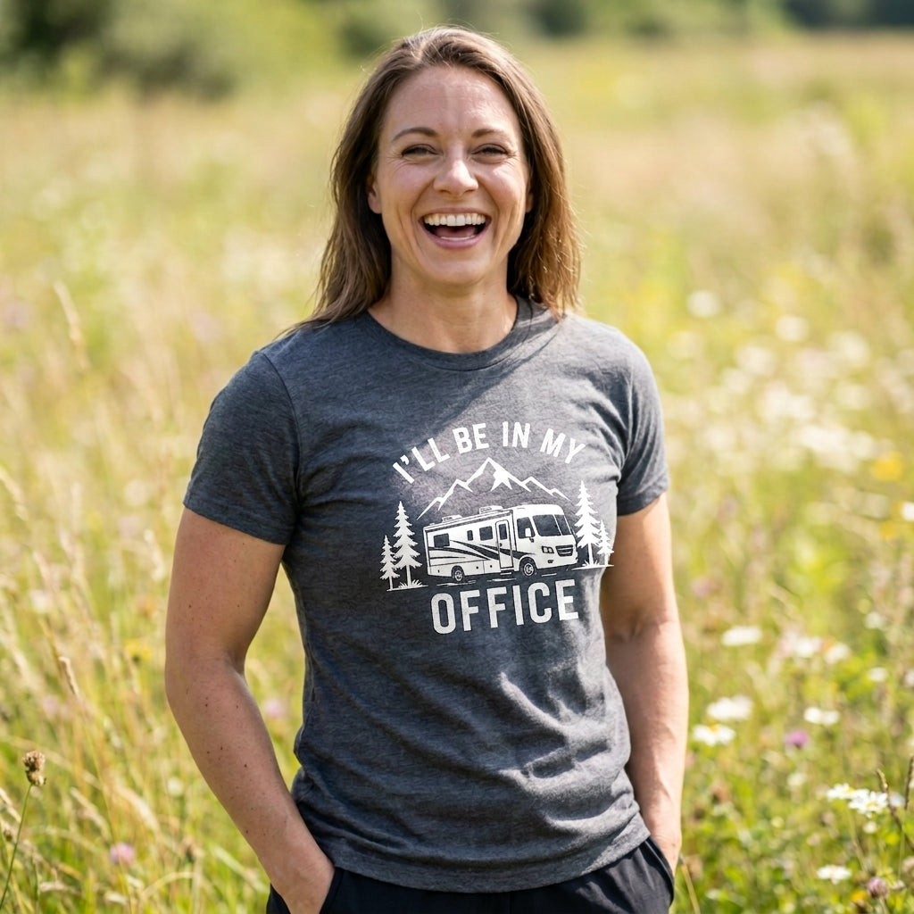 A woman smiling in a grassy field, wearing a dark heather gray t-shirt with a white graphic that reads 'I'LL BE IN MY OFFICE' with an RV and mountains.