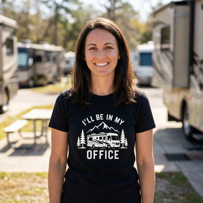 A smiling woman stands outdoors in a campground or RV park, wearing a black short-sleeve T-shirt with a white graphic that reads 'I'LL BE IN MY OFFICE' above and below an RV and pine tree illustration.