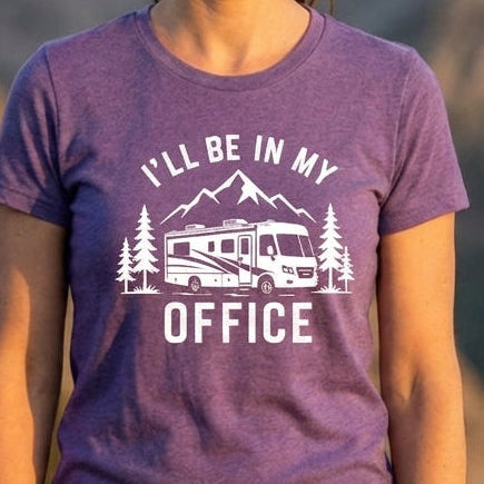 A woman wearing a purple graphic t-shirt with a white RV and mountain design, standing in front of a blurred mountain landscape at sunset.
