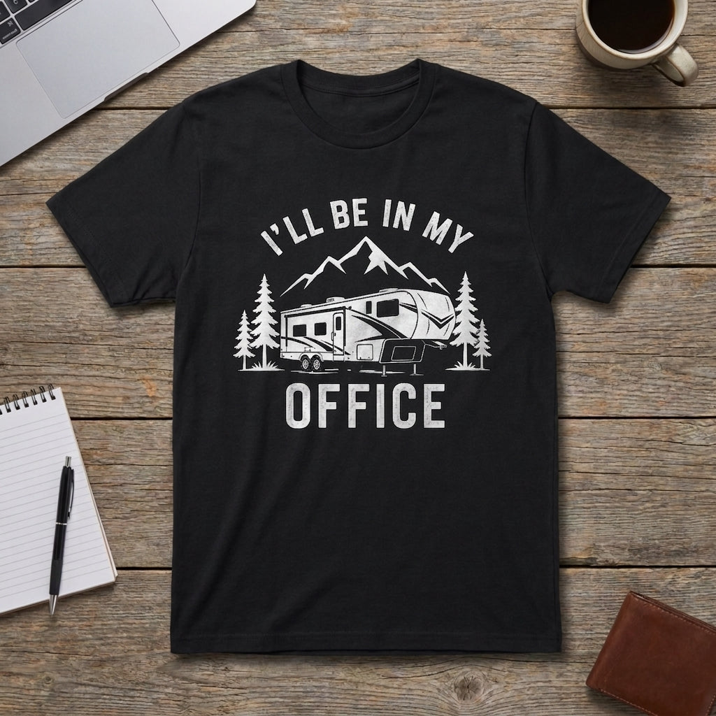 Black I'll Be In My Office fifth wheel t-shirt laid flat on a wooden table with a laptop, coffee mug, notebook, pen, and wallet, showing an RV, mountains, and pine trees graphic.