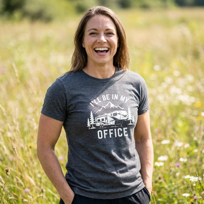 Smiling woman in a dark heather gray I'll Be In My Office fifth wheel t-shirt with a camper and mountains graphic, standing in a grassy field.
