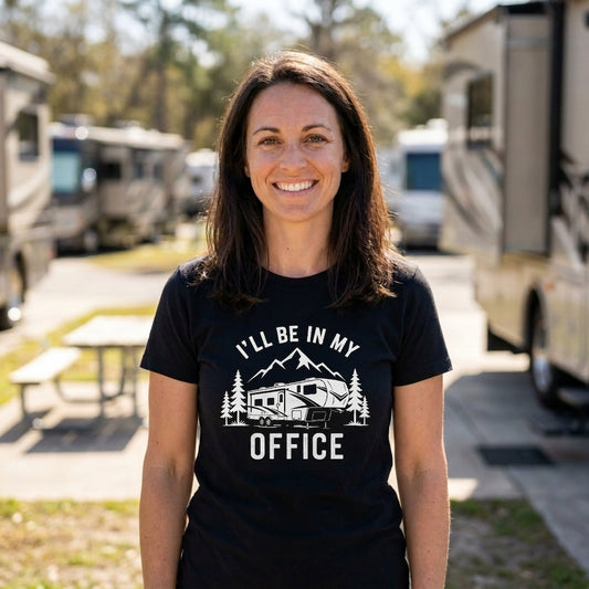 A smiling woman stands outdoors in a campground or RV park, wearing a black short-sleeve T-shirt with a white graphic that reads 'I'LL BE IN MY OFFICE' above and below an RV and pine tree illustration.