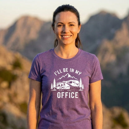 Woman in a purple I'll Be In My Office fifth wheel t-shirt with a white fifth wheel and mountain design, standing in front of a mountain landscape at sunset.