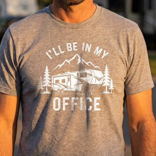 A man wearing a heather gray t-shirt with a white graphic that reads 'I'LL BE IN MY OFFICE' with a camper and mountains, standing outdoors in front of blurred RVs at sunset.