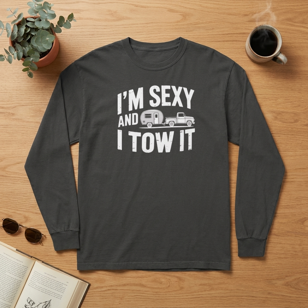 Flat lay of a charcoal gray I'm Sexy and I Tow It long sleeve shirt on a wood table with a coffee mug, sunglasses, a plant and an open book
