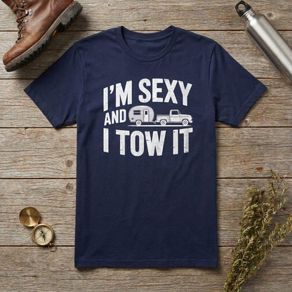 A navy blue crewneck t-shirt with a white graphic print reading 'I'M SEXY AND I TOW IT' with a trailer and truck illustration, laid flat on a rustic wooden surface with outdoor props including a boot, compass, bottle, and dried flowers.