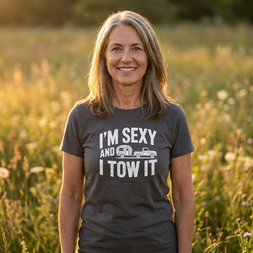 A smiling woman standing in a sunlit grassy field wearing a dark charcoal gray graphic t-shirt with the white text 'I'M SEXY AND I TOW IT' and a small towing truck and RV illustration.