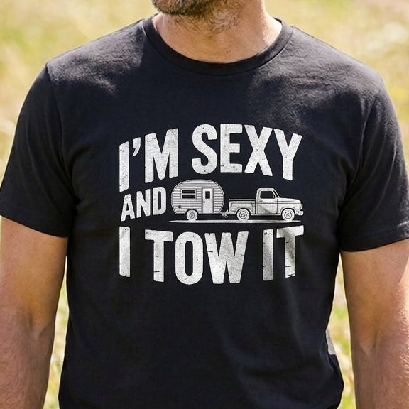 A man wearing a black graphic t-shirt with the white text 'I'M SEXY AND I TOW IT' and a small truck and camper graphic, standing in a sunny grassy field.