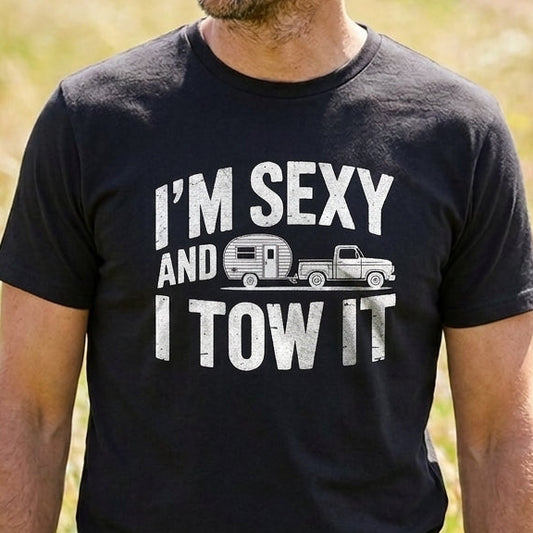 A man wearing a black graphic t-shirt with the white text 'I'M SEXY AND I TOW IT' and a small truck and camper graphic, standing in a sunny grassy field.