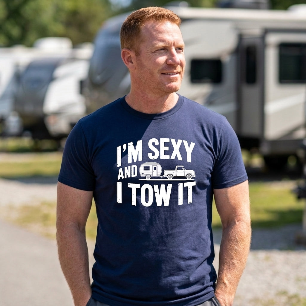 A man wearing a navy blue graphic t-shirt that reads 'I'M SEXY AND I TOW IT' with a small truck and camper trailer illustration, standing outdoors in front of blurred RVs.