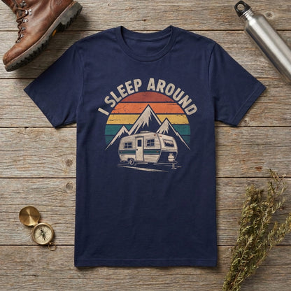 Navy blue I Sleep Around t-shirt with a retro camper and mountains graphic laid flat on a wooden surface with a boot, water bottle, compass, and dried flowers.